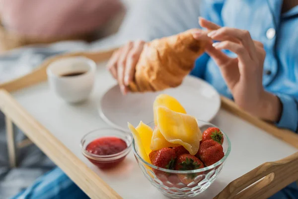 ベッドで朝食をとりながらジャムとドライマンゴーをトレイの近くに新鮮なクロワッサンを持っている女性のクロップドビュー — ストック写真