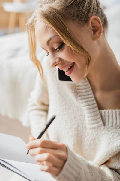 穿着毛衣笑着的女人在智能手机上说话 在笔记本上记笔记 — 图库照片