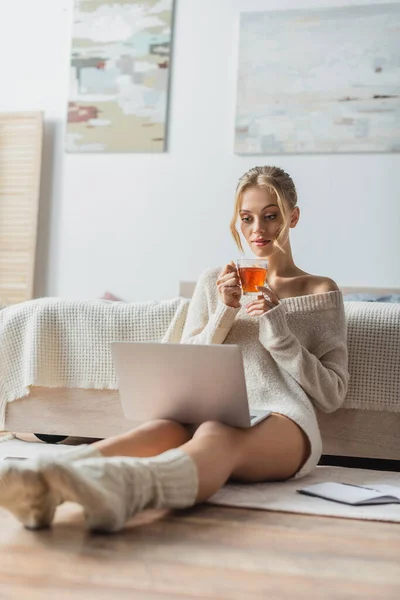 在现代公寓里 金发女人一边拿着杯子喝茶 一边看笔记本电脑 — 图库照片