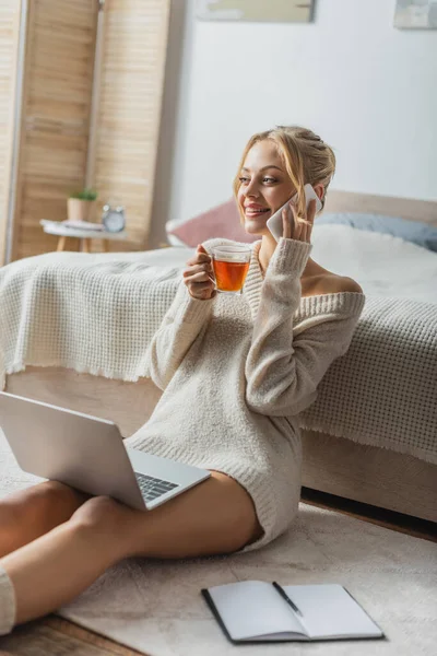 ラップトップに座って現代のアパートでスマートフォンで話をしながらお茶を飲みながら幸せな女性 — ストック写真