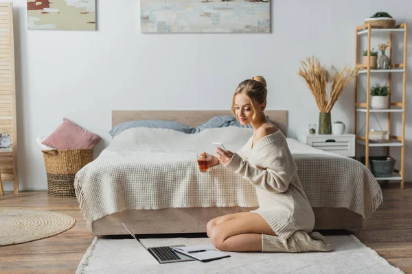 金发女人端着杯子喝茶 在靠近笔记本电脑的地方用智能手机在地毯上 — 图库照片