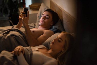 unfaithful man using mobile phone and looking at blonde girlfriend sleeping in bed, cheating concept  clipart