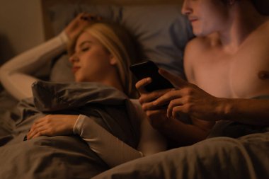 Üzgün adam yatakta uyuyan sarışın kız arkadaşının akıllı telefonunu kontrol ediyor, aldatma konsepti 