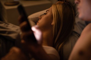 blonde woman sleeping next to unfaithful man with smartphone, cheating concept  clipart