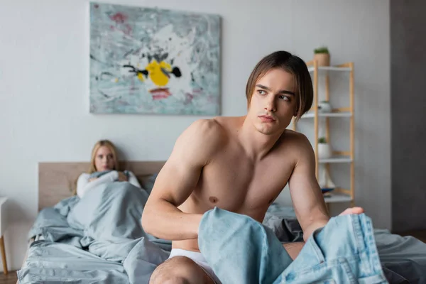 Mężczyzna Bez Koszulki Trzyma Jeansy Siedząc Łóżku Pobliżu Rozmytej Kobiety — Zdjęcie stockowe