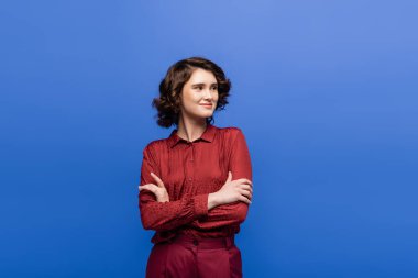 Kısa kıvırcık saçlı, kolları katlanmış, mavi renkli neşeli bir kadın. 