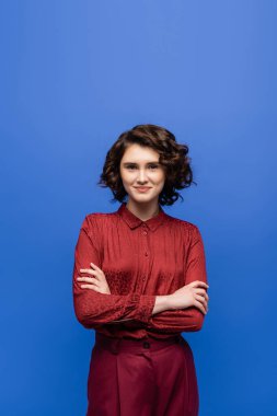 Kısa kıvırcık saçlı neşeli bir öğretmen kollarını katlamış mavinin üzerinde duruyor. 
