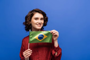 happy language teacher smiling while holding flag of Brazil isolated on blue  clipart