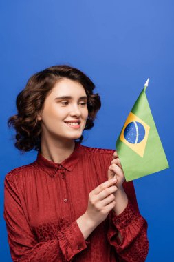 cheerful language teacher smiling while holding flag of Brazil isolated on blue  clipart