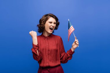 Amerika bayrağı taşıyan neşeli genç bir kadın mavi üzerine izole edilmiş sevinçle seviniyor.