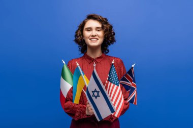 Kameraya gülümseyen memnun bir kadın, elinde mavi üzerine izole edilmiş farklı ülkelerin bayraklarıyla