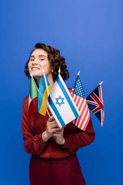 Genç, ilham verici bir kadın, elinde mavi üzerine izole edilmiş farklı ülkelerin bayraklarını tutarken kameraya bakıyor.
