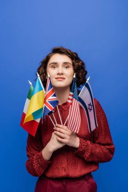 Çeşitli ülkelerin bayraklarını taşıyan hayret verici bir kadın ve mavi renkli kameraya bakıyor.