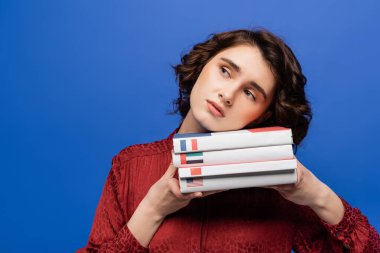 bored student holding textbooks of foreign languages while looking away isolated on blue clipart