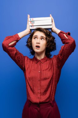 shocked student opening mouth and holding textbooks of foreign languages over head isolated on blue clipart