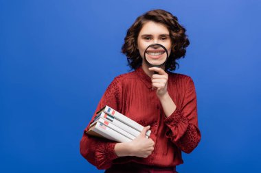 Yabancı diller hakkında ders kitapları olan neşeli öğrenci maviye izole edilmiş büyüteçle eğleniyor ve gülümsüyor.
