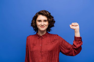 young joyful woman in red blouse success gesture while smiling at camera isolated on blue clipart