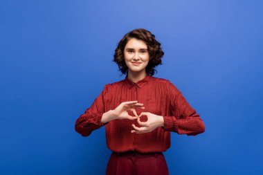 Genç ve neşeli bir kadın kameraya bakıyor ve mavi üzerine izole edilmiş işaret dilinde tercüman anlamına geldiğini gösteriyor.
