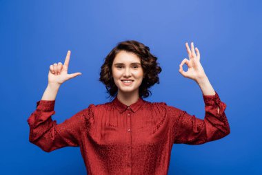 pleased teacher smiling at camera and showing alphabet letters on sign language isolated on blue clipart