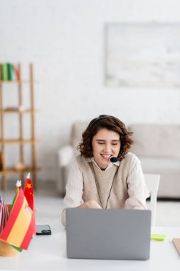 Evde akıllı telefon ve uluslararası bayrakların yanındaki dizüstü bilgisayarla çevrimiçi derste konuşan kaygısız dil öğretmeni. 