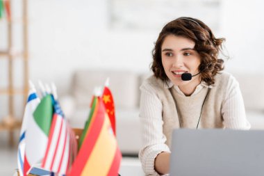 Bilgisayarın yanında online ders sırasında gülümseyen kulaklıklı genç dil öğretmeni ve evdeki uluslararası bayraklar