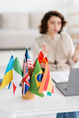 Bulanık dizüstü bilgisayarın yanındaki uluslararası bayrakların seçici odak noktası ve evde çalışan genç dil öğretmeni