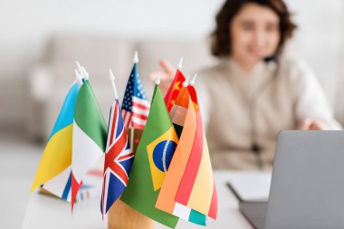 Bulanık dizüstü bilgisayarın yanında çeşitli uluslararası bayrakların seçici odak noktası ve evde online ders alan dil öğretmeni