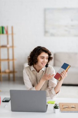 Evde online ders sırasında bilgisayarın yanında Fransızca ders kitabına işaret eden kulaklık takan gülen dil öğretmeni.