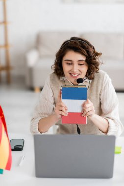 Evde dizüstü bilgisayar dersi sırasında Fransızca ders kitabı gösteren kulaklıklı neşeli öğretmen.