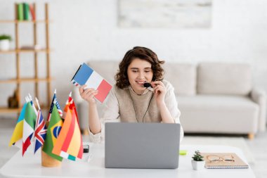 Online dizüstü bilgisayar dersi sırasında uluslararası bayrakların yanında Fransızca ders kitabı tutan kulaklıklı mutlu dil öğretmeni 