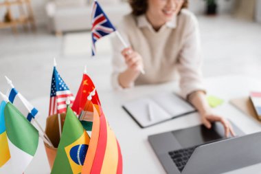 Bulanık arkaplanda dizüstü bilgisayar kullanarak yabancı dil öğretmeninin yanındaki uluslararası bayrakların seçici odak noktası 