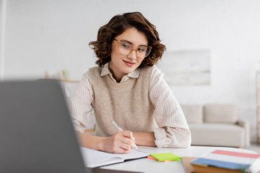smiling woman in glasses taking notes while learning foreign language and watching online lesson on laptop  clipart