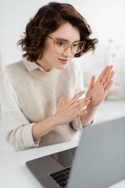 cheerful teacher in glasses showing two handed sign language gesture while looking at laptop  clipart