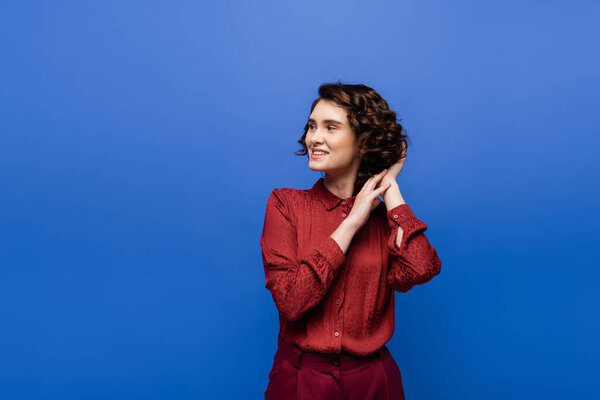 довольный учитель языка в красной блузке регулируя кудрявые волосы и глядя в сторону изолированы на синий 