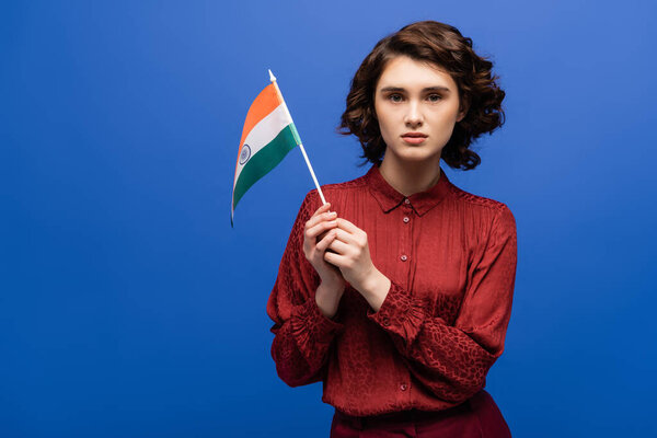 curly language teacher holding flag of India and looking at camera isolated on blue 