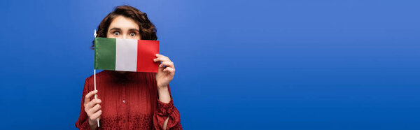 young curly woman covering face while holding flag of Italy and looking at camera isolated on blue, banner  