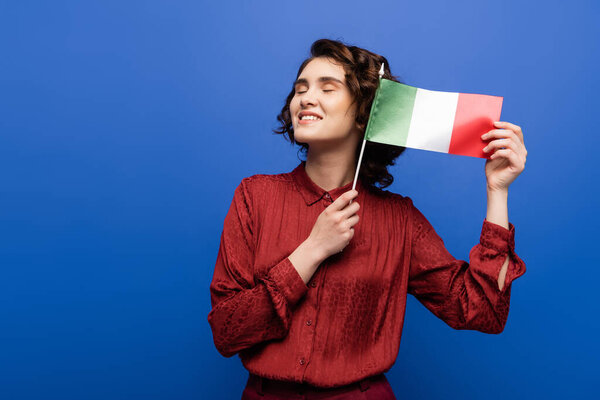 pleased language teacher smiling while holding flag of Italy isolated on blue 