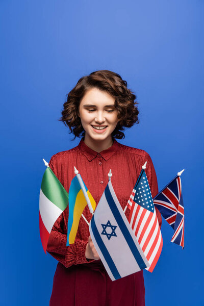 happy woman with wavy brunette hair looking at international flags isolated on blue