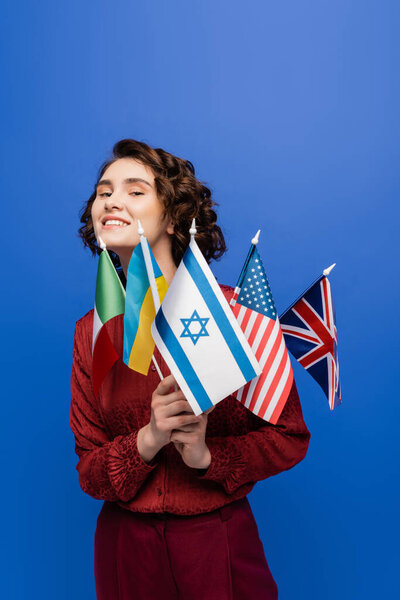 young inspired woman looking at camera while holding flags of different countries isolated on blue
