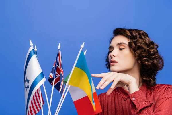 brunette woman pointing at different flags while choosing language to study isolated on blue
