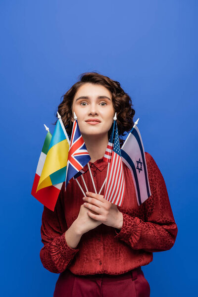 amazed woman holding flags of various countries and looking at camera isolated on blue