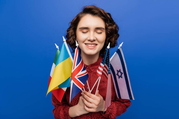 inspired woman with closed eyes holding flags of different countries isolated on blue