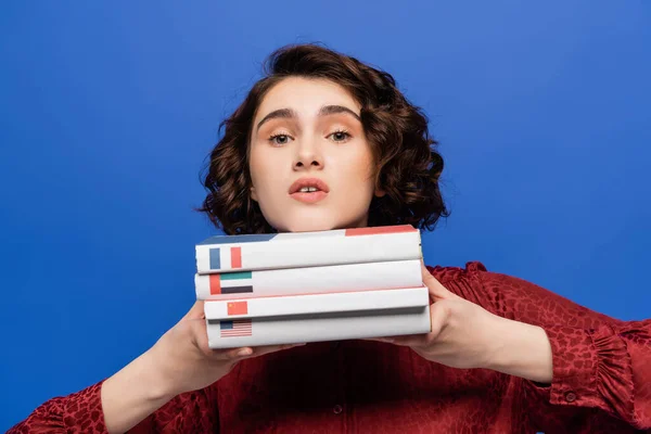 Stock image discouraged and tired student looking at camera while holding textbooks of foreign languages isolated on blue