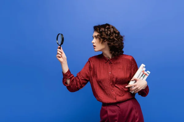 attentive student holding textbooks of foreign languages and looking through magnifier isolated on blue