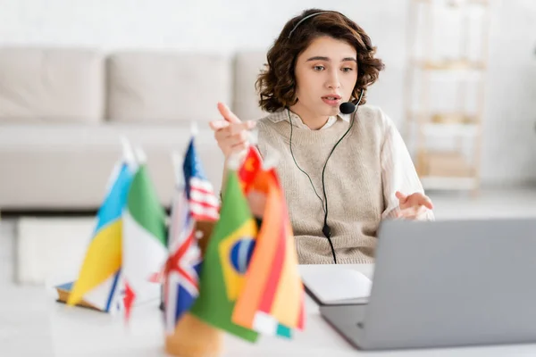 stock image language teacher in headset pointing with fingers while talking during video chat on laptop near blurred flags at home