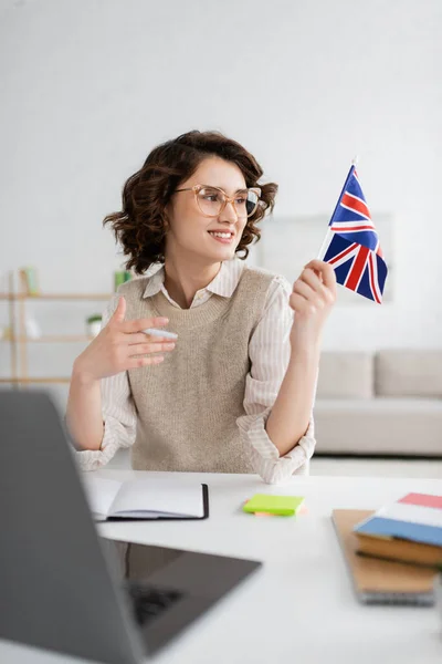 英国の国旗を見ながら家でぼやけたノートパソコンの近くに座っている陽気な語学教師 — ストック写真