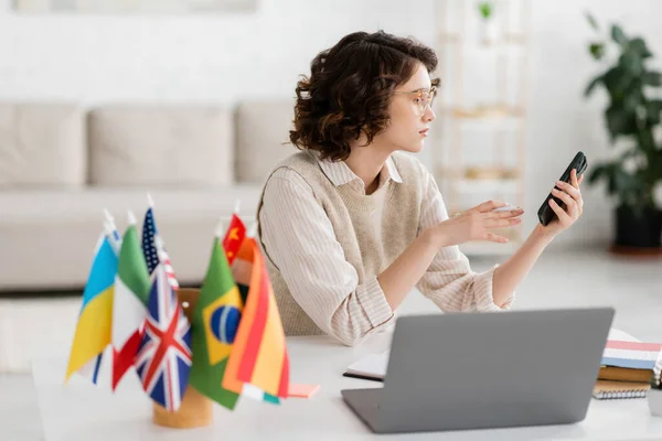 Young Language Teacher Glasses Holding Smartphone Laptop International Flags Blurred — Stock Photo, Image