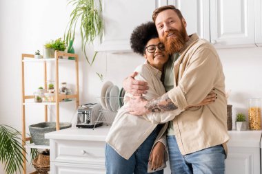 Gülümseyen sakallı adam mutfakta dururken gözlüklü Afrikalı Amerikalı kız arkadaşına sarılıyor.  