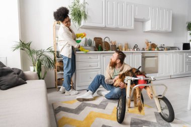 Neşeli Afro-Amerikalı kadın, erkek arkadaşının yanında mutfağı temizliyor ve tekerlekli sandalyede sakat bir köpek var. 
