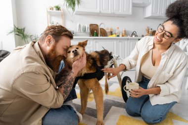 Gülümseyen Afrikalı Amerikalı kadın elinde kahve, erkek arkadaşı da engelli köpeğini okşuyor. 